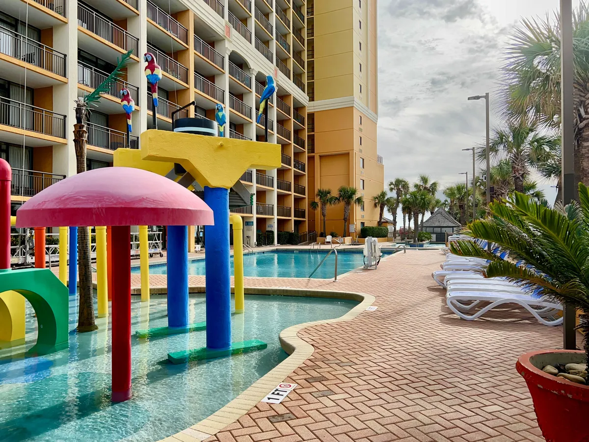 A lively outdoor pool area featuring colorful water play structures, inviting swimming pools, sun loungers, and palm trees. Families and children are actively enjoying the water, creating an atmosphere of fun and relaxation. The scene is bright, playful, and clearly designed for both excitement and comfort.