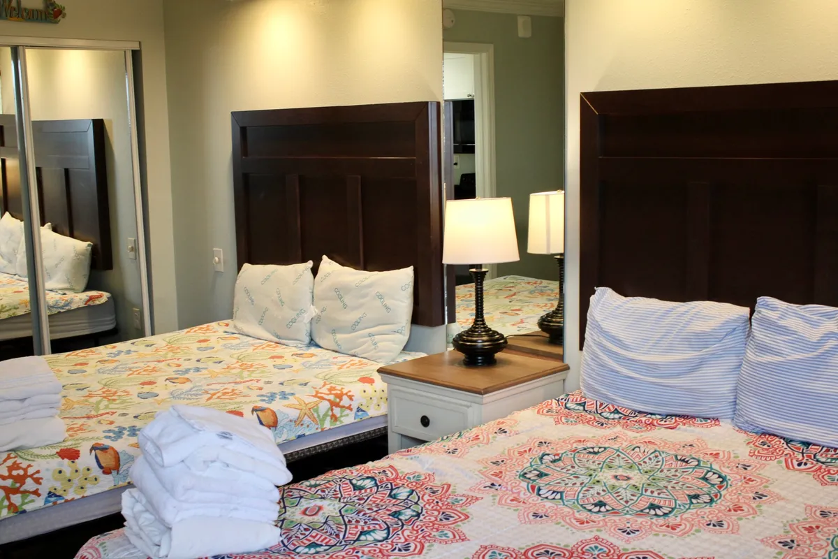 The image showcases the condo's inviting bedroom, featuring two queen beds with plush bedding, soft natural sunlight filtering through the windows, warm lighting from bedside lamps, and mirrors that enhance the room's spacious feel. The overall atmosphere is cozy, bright, and perfect for unwinding after a day at the beach.