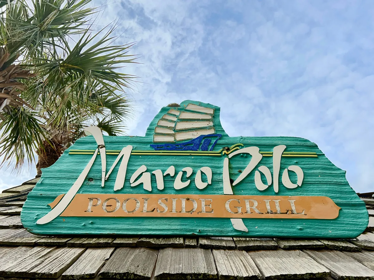 The exterior features the Marco Polo Poolside Grill signage surrounded by palm trees and a clear blue sky, suggesting an inviting spot for casual outdoor dining and socializing. The setting feels relaxed and distinctly vacation-like, perfect for leisurely meals or sunset drinks steps from the ocean.