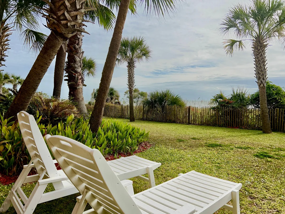 An exterior lounging area with white lounge chairs arranged around lush palm trees and fenced greenery, all beneath a wide sky with clouds. The space is inviting for reading, relaxing, or casual socializing, with a sense of privacy and tranquility.