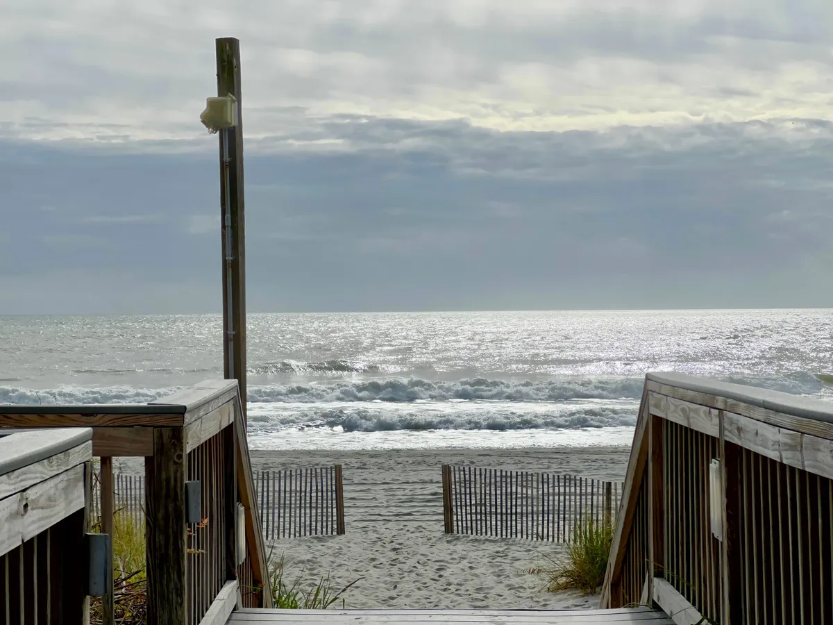 A wooden walkway leads directly toward the sandy beach, with gentle ocean waves in the background and a beach fence guiding the path. The sky is slightly cloudy, creating a welcoming yet active beach atmosphere.