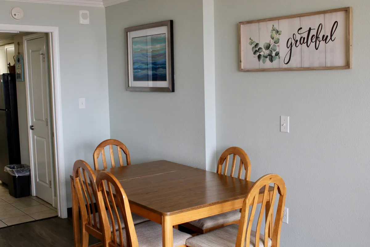 A cozy dining area with a wooden dining table surrounded by matching chairs, adorned with framed artwork and a welcoming decorative sign. The space is well-lit and designed for sharing meals, playing games, or gathering for casual conversation, embodying a sense of togetherness and warmth.
