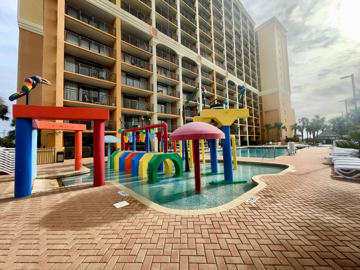 A vibrant pool complex featuring colorful water play structures, a swimming pool, tropical plants, and building balconies visible in the background. Children are playing and splashing in the water, while adults relax poolside, creating a lively and family-friendly atmosphere.