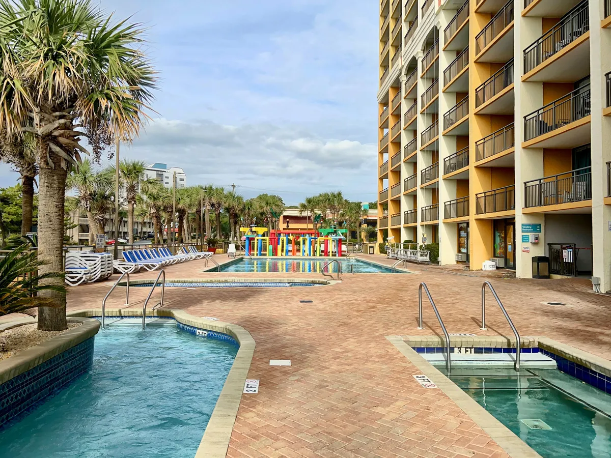 A cheerful and relaxed outdoor scene with a swimming pool, a colorful water play area filled with children, palm trees, and the resort’s balconies overlooking the space. The setting is lively with plenty of space for family gatherings and relaxation.
