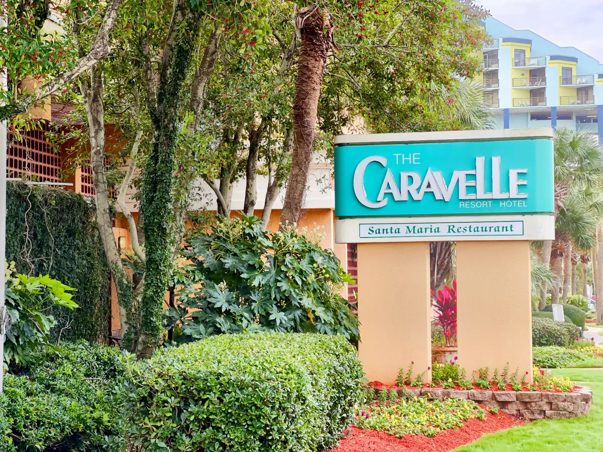 The welcoming entrance to The Caravelle Resort featuring its distinctive hotel sign, lush green landscaping, and a mention of the Santa Maria Restaurant. The scene hints at the broader amenities and vibrant local area available for exploration.
