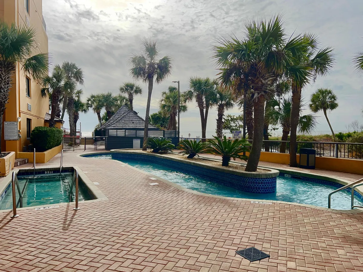 This image features an expansive outdoor swimming pool surrounded by palm trees, a hot tub, a lounge area, and a cabana. The pool area is designed for both relaxation and socializing, offering plenty of space for groups to gather. The setting captures a leisurely mood with the ocean visible in the background, evoking a sense of vacation and fun.