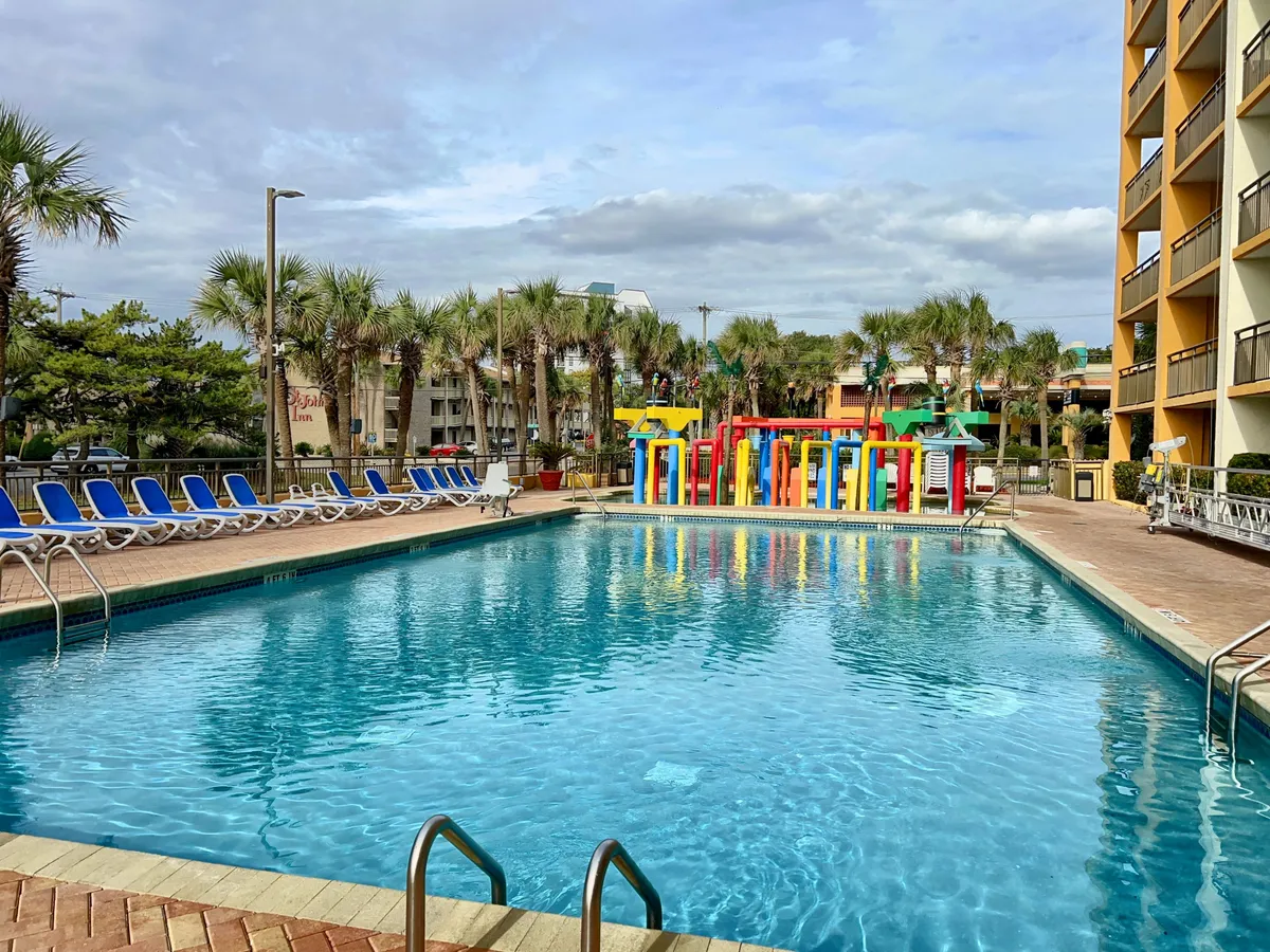 A cheerful pool scene featuring a spacious swimming pool, colorful play structures, sun loungers, and palm trees. Families and friends are pictured enjoying the water, sunbathing, and socializing. The vibrant atmosphere suggests an environment where group fun and playful moments are always within reach.