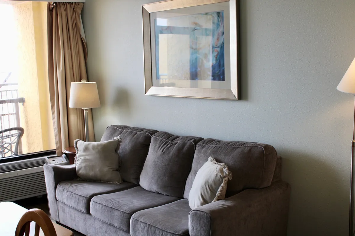 A bright and comfortable living area featuring a plush sofa, decorative pillows, framed artwork, and a modern floor lamp. The room is arranged for relaxation and socializing, with inviting seating that encourages conversation or enjoying a quiet moment with coffee. Large windows allow natural light to bathe the space, contributing to its airy ambiance.
